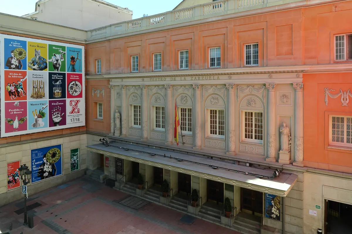 La fachada de piedra rosa y crema del Teatro de la Zarzuela, vista desde un patio con carteles en una pared.