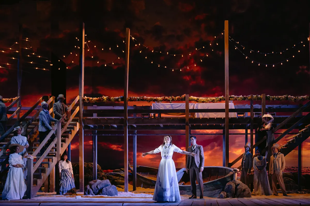 Dos cantantes sobre un decorado con un cielo de atardecer rojo y dorado, un embarcadero de madera y guirnaldas de luces.