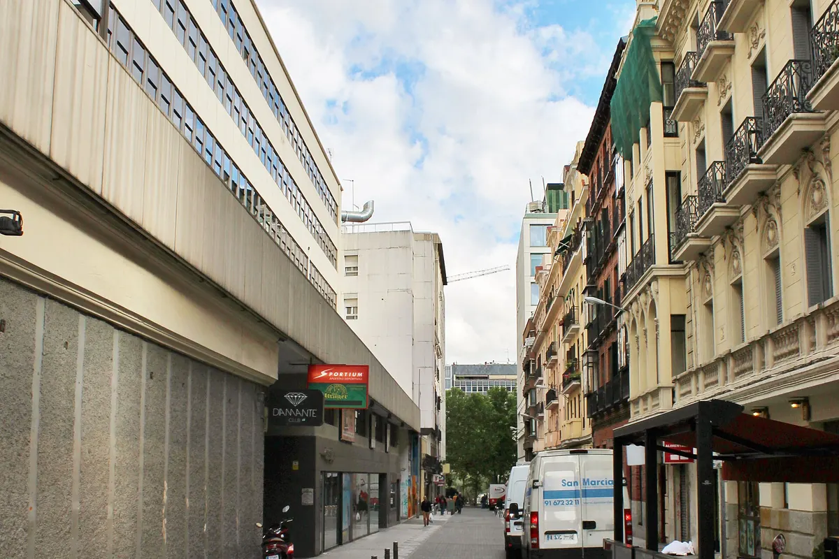 Una tranquila calle urbana con un largo edificio moderno a la izquierda, bloques de viviendas color crema con balcones a la derecha y un cielo nublado